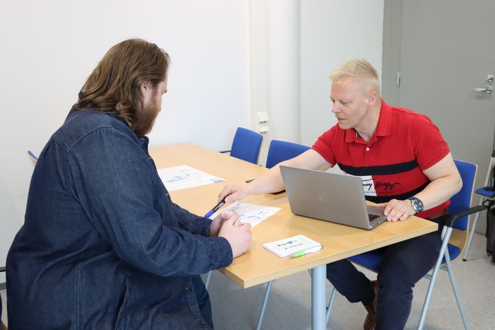 Nuorten start up -pilotin projektipäällikkö Tero Eklund istuu tuolilla pöydän ääressä pilotin asiakkaan kanssa. Terolla on tietokone auki ja pöydällä on papereita.