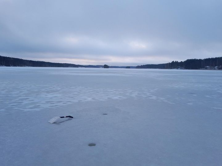 Laaja, lumen peittämä jäätyneen Yöveden pinta pilvisen taivaan alla. Ympäröivä maisema sisältää kaukaiset, puurajaiset rannat harmaan, pilvisen taivaan alla. Ilmapiiri vaikuttaa kylmältä ja rauhalliselta. Jäällä lumilapio ja kaksi reikää