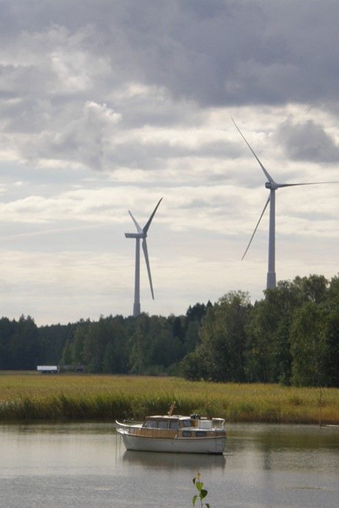 Kaksi tuulivoimalaa taivasta vasten eteläsavolaisessa maisemassa. Etualalla kulkee pieni laiva järvellä.