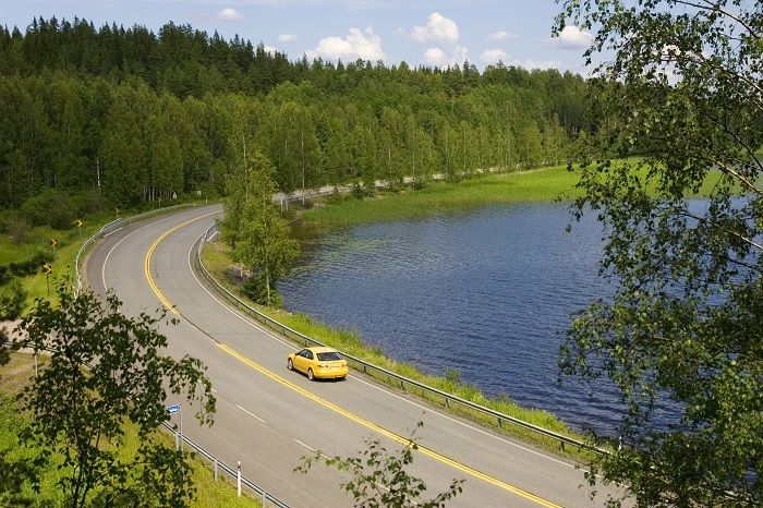 Keltainen auto ajamassa kaarteeseen kesäisessä maisemassa järven rannalla.