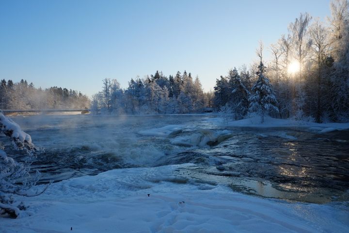 Talvinen jokimaisema auringon paistaessa puiden takaa.