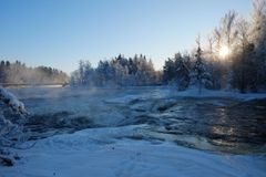 En flod omgiven av snötäckta träd i vinterlandskap, med soluppgång i bakgrunden.