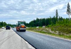 Tiehöyryjyrä tasoittaa juuri päällystettyä tietä, ympärillä on metsämaisemaa ja muutamia autoja.