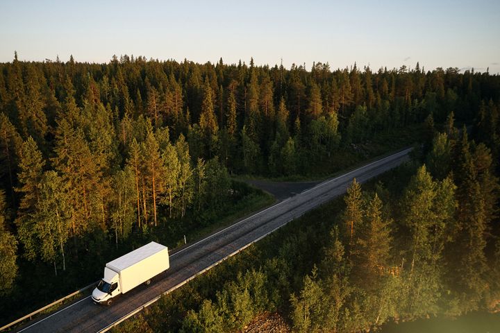 Valkoinen kuorma-auto ajaa pitkää tietä metsän keskellä.