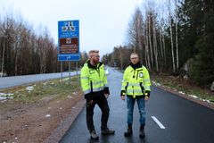 Två personer i reflexjackor står på en asfalterad väg nära en skylt.