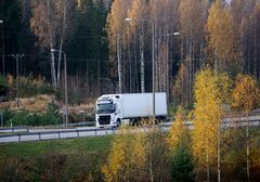 Valkoinen kuorma-auto ajaa syksyisessä maisemassa, taustalla näkyy keltaisia ja ruskeita puiden lehtiä.