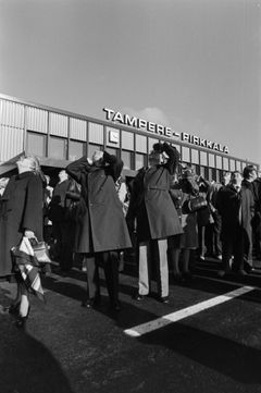 Ihmisiä seisoo ulkona Tampere-Pirkkalan lentoaseman avajaisissa vuonna 1979, katsoen yläpuolelle. Taustalla näkyy lentoaseman rakennus.