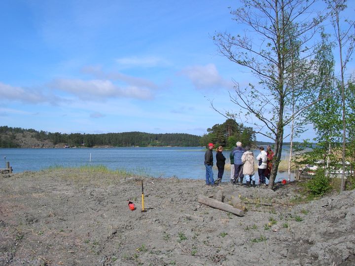 Joukko ihmisiä kokoustaa järven rannalla ja maahan on jo puoliksi kiinnitetty rajapyykki