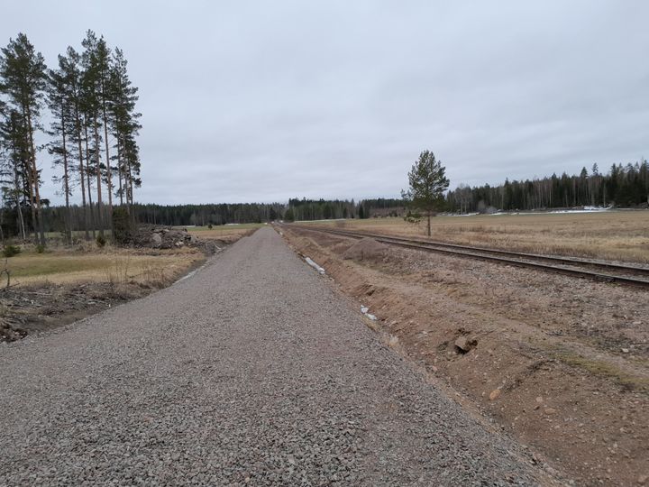 Kuvan keskellä näkyy eteenpäin kulkeva hiekkatie. Hiekkatien oikealla puolella on junarata. Molemmissa reunoissa näkyy peltoa ja puita.