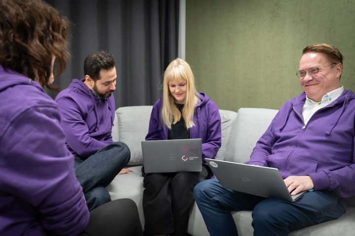 Kuvassa on neljä Maanmittauslaitoksen työntekijää, joilla on violetit hupparit. Yhdellä on sylissä kannettava tietokone.