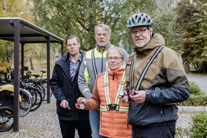 Kolme miestä ja yksi nainen seisoo ulkona pyörätelineiden edustalla. Yhdellä miehistä on päässään pyöräilykypärä, kaikilla on asussaan  erilaisia heijastimia.