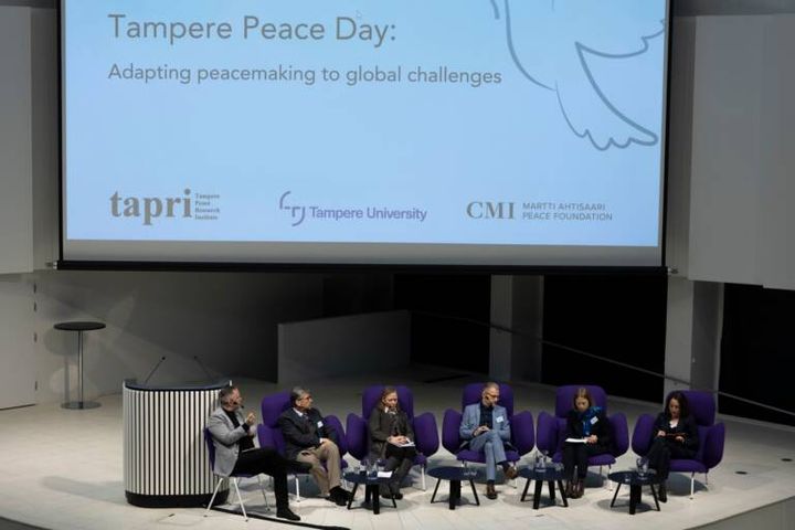 Paneelikeskustelu Tampere Peace Day -tapahtumassa. Kuusi henkilöä istuu lavalla keskustelemassa. Taustalla tapahtuman nimi ja organisaatioiden logot.