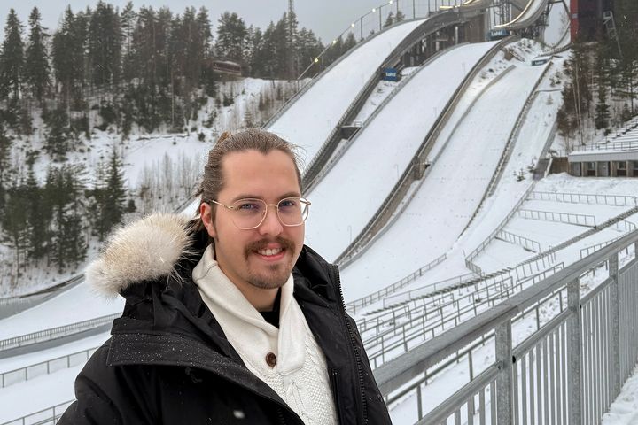 Henkilö hymyilee lumisessa maisemassa hyppyrimäkien edessä.