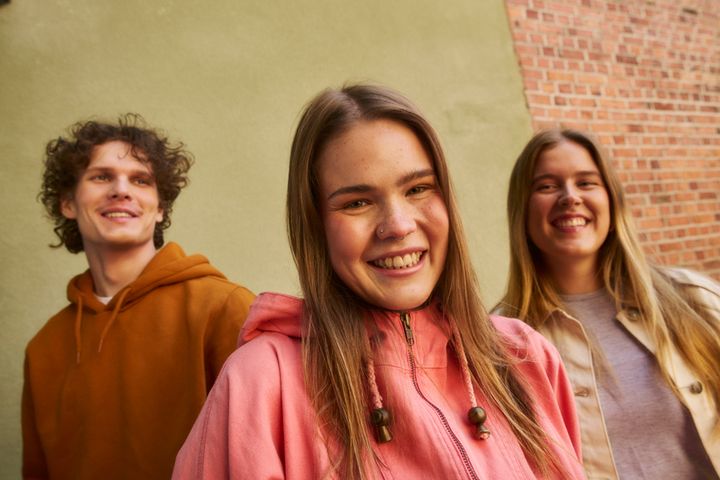 Kolme hymyilevää henkilöä värikkäissä vaatteissa seisoo ulkona tiiliseinän edessä.