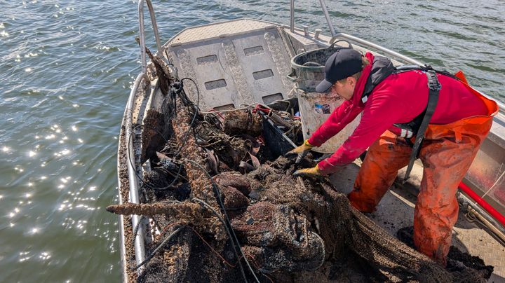 En man klädd i röd huvtröja och orange vadarbyxor lyfter fisknät från en hög med nät och annat lerigt skräp på botten av båten.