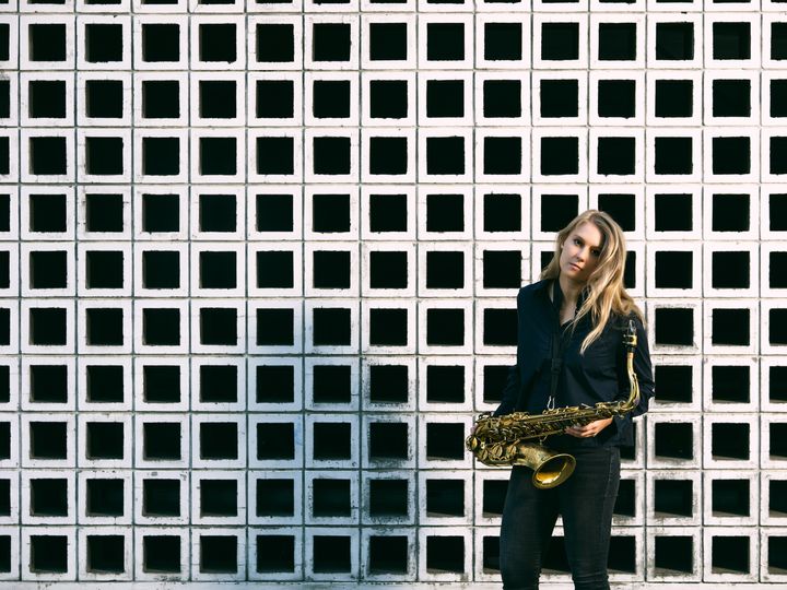 Saxophonist Adele Sauros stands with her saxophone in front of a black-and-white checkered background.