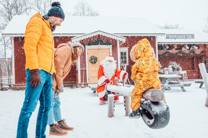 Perhe viettää aikaa lumisateessa ulkona, lapsi keinuu ja joulupukki istuu taustalla.