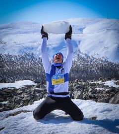 Talviasuun pukeutunut henkilö polvistuneena lumipeitteisellä vuorella ja nostaa suuren jäälohkareen päänsä yläpuolelle. Taustalla lumisia maisemia.