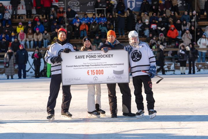 Neljä henkilöä seisoo jäällä pidellen 15 000 euron lahjoitustsekkiä Luonnonperintösäätiölle Save Pond Hockey -turnauksessa. Taustalla näkyy katsojia.
