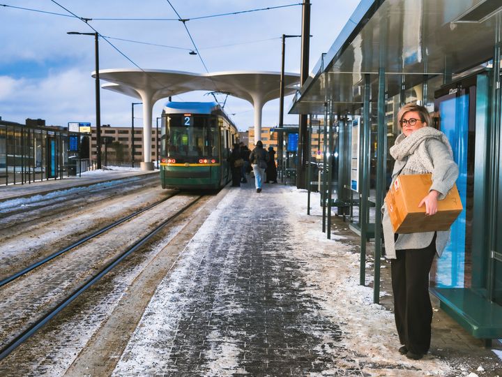 Nainen odottaa raitiovaunua lumisella pysäkillä. Hänellä on iso paketti sylissään.