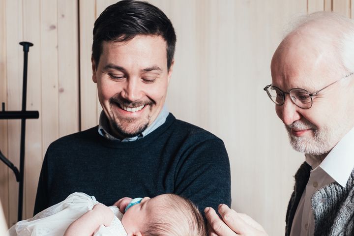 En ung pappa håller leende sitt barn i dopklänning i famnen och en äldre släkting, mor- eller farförälder står bredvid.