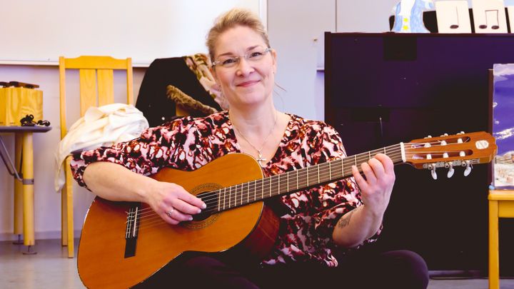 En kvinna i glasögon och upsatt hår sitter på golvet med en gitarr i sin famn.
