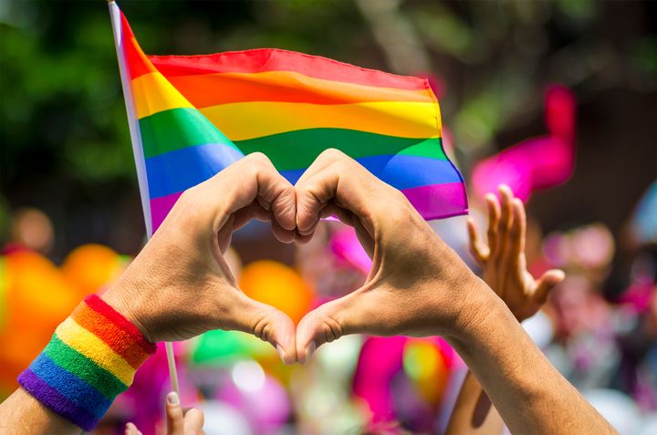 Ihmisjoukko Pride-tunnelmissa. Etualalla oleva henkilö muodostaa käsillään sydämen. Taustalla joukko ihmisiä ja värikäs sateenkaarilippu.