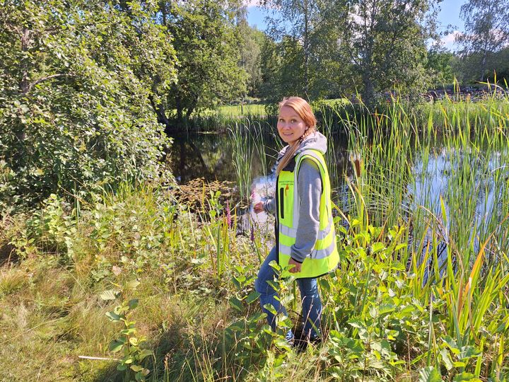 Viia Löppönen står i gul arbetsväst intill strandvegetation som växer intill en sjö.