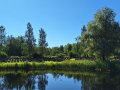 Naturskönt landskap där gravstenar och lummiga träd skymtar bakom en spegelblank sjö i förgrunden.