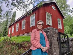 En kvinna i rosa täckjacka lutar sig mot ett räcke och i bakgrunden den röda gamla sockenstugan i trä intill Esbo domkyrka.