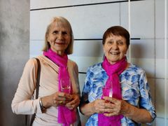 Två kvinnor står med champagneglas i händerna och iklädda starkt rosa långa scarfar.