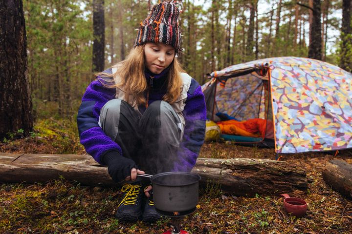 En ung kvinna eller flicka sitter i skogen intill en lägereld och i bakgrunden syns ett tält.