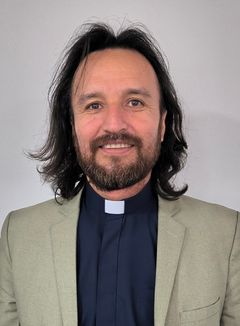 En man med mörkt halvlångt hår och iklädd prästkrage och kavaj.