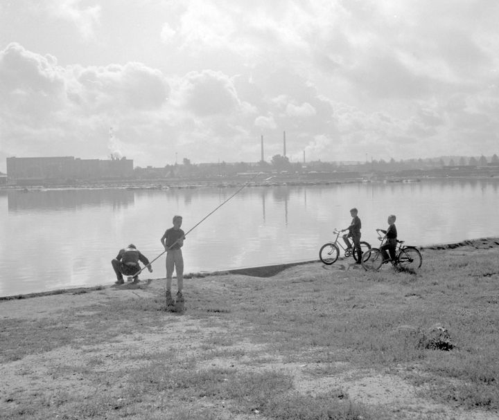 Mustavalkoinen valokuva: ihmisiä kalastamassa ja kaksi henkilöä pyörillä Ratinan suvannolla, taustalla teollisuusrakennuksia, vuosi 1956.