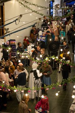 Alt text: Ihmiset kerääntyneet myyntikojuille taidekujan sisätiloissa, joissa on ripustettu koristevaloja ja -köynnöksiä.