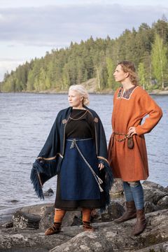 Kaksi henkilöä muinaispuvuissa seisoo veden äärellä, taustalla metsä.