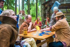 Loveroolipelaajat istuvat puisen pöydän ympärillä rooliasuissa ja pelaavat korttipeliä. Ympärillä katselijoita. Taustalla metsää ja puinen rakennus.