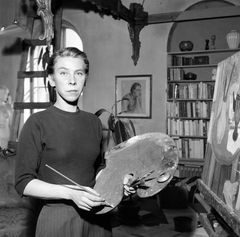 Mustavalkoinen vanha valokuva, jossa nainen seisoo huoneessa maalauspaletti kädessään.