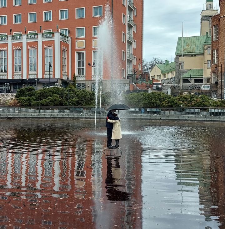 Punaisen rakennuksen edessä on lampi, jossa on suihkulähde. Suihkulähteen vieressä seisoo realistisen näköinen pariskunta sateenvarjon alla.