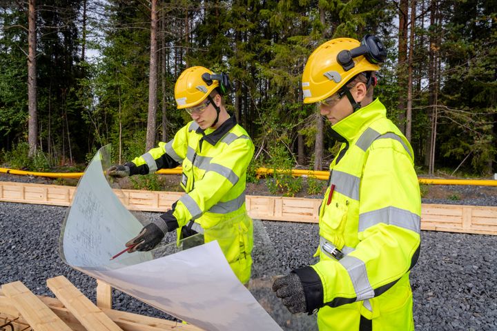 Kaksi talnrakentajaa tarkastelevat rakennuspiirustuksia. Miehillä on työasut päällä. Taustalla kesäinen mäntymetsä.