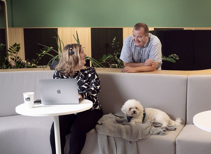 En person med laptop och hörlurar sitter på en soffa och pratar med en stående man. En vit hund ligger bredvid på soffan.