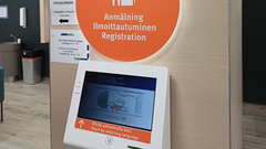 Kuvan ylälaidassa näkyy seinällä iso oranssi kyltti, jossa lukee suomeksi, ruotsiksi ja englanniksi ilmoittautuminen. Kyltin alla on ilmoittautumisautomaatin näyttöruutu.