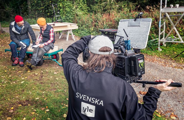 Kuvassa on etualalla selin tummassa Svenska Yle -takissa kuvaaja, joka kuvaa kahta ihmistä istumassa ulkosalla.