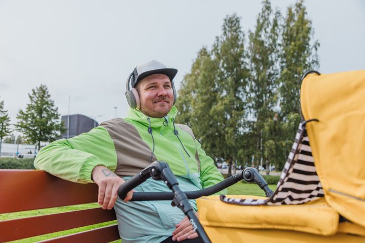 Mies istuu hymyillen puistonpenkillä kuulokkeet päässä ja työntelee keltaisia lastenvaunuja.