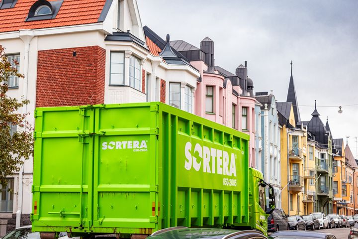 Vihreä jätekuljetusauto, jossa Sorteran nimi isolla kyljessä ajaa värikkäiden kerrostalojen edessä.