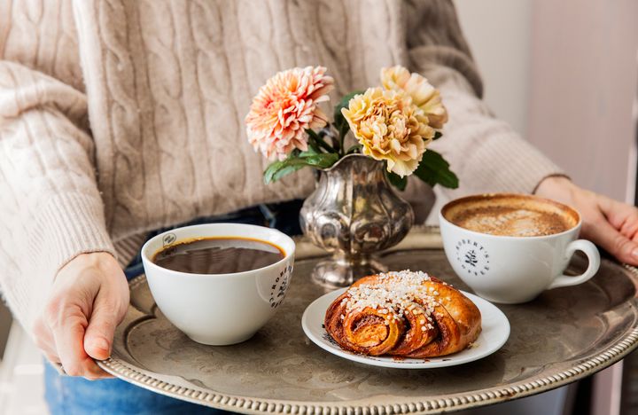 Henkilö pitelee tarjotinta, jossa on kahvikuppi, cappuccinokuppi, korvapuusti ja maljakossa kukkia.