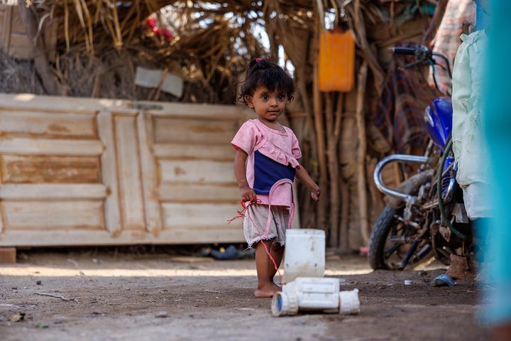 Pieni lapsi seisoo hiekkapihalla vaaleanpunaisessa paidassa leikkien vaalealla leluautolla. Taustalla näkyy yksinkertaisia rakennuksia ja moottoripyörä.