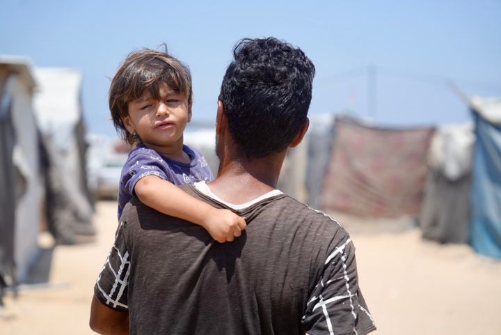 Mies kantaa pientä lasta olkapäillään leirillä olevien telttojen taustalla.