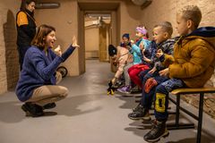 Opettaja kommunikoi lasten kanssa, kun he istuvat penkillä sisätiloissa väestönsuojassa talvivaatteet päällään.