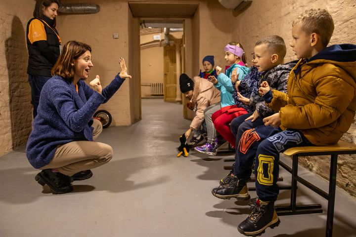 Opettaja kommunikoi lasten kanssa, kun he istuvat penkillä sisätiloissa väestönsuojassa talvivaatteet päällään.